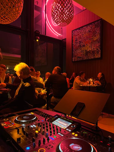 The image captures a dimly lit, vibrant restaurant setting. A group of people are seated around a table, enjoying food and drinks. The interior is illuminated with pink neon lights and decorative wicker lamps. A piece of modern art hangs on the wall. A DJ setup is prominently featured in the foreground, suggesting live entertainment is part of the restaurant's atmosphere. The overall ambiance suggests a lively and stylish dining experience.