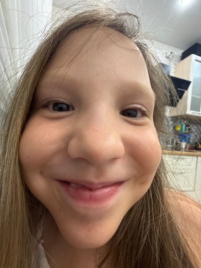 The image is a close-up, distorted selfie of a young girl with long brown hair. Her face is the primary subject, filling most of the frame. She has brown eyes, a wide smile revealing some teeth, and her cheeks appear to be puffed out. The background is blurry, showing what appears to be an indoor setting with cabinetry and possibly curtains. There is no visible indication that this image was taken in Shushary, Russia.