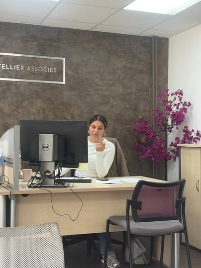 The image displays an indoor office environment, identified as TELLIER ASSOCIES, located in Cagnes-sur-Mer, France. A female individual with dark hair, wearing a white long-sleeved top, blue jeans, and metallic athletic shoes, is seated at a light wood-colored desk. She is looking directly at the camera with her right hand raised, displaying her extended middle finger.
On the desk, a black Dell computer monitor with a visible Dell logo is centrally positioned, with a computer tower mounted behind it. To the left of the monitor, a pink mug, a document holder, and some papers are present. To the right, additional papers and a dark-colored smartphone are visible.
Behind the seated individual, the wall features a dark brown, textured finish, upon which a silver-framed sign reads "TELLIER ASSOCIES". To the right of the individual, a large artificial plant with numerous purple flowers stands next to a light wood-colored two-door cabinet. The wall to the far right is white, with vertical pipes running along the corner. A purple mesh-backed office chair is positioned in the mid-ground, facing away from the viewer. A portion of another light-colored mesh-backed office chair is visible in the foreground to the left. The ceiling is composed of white acoustic tiles with recessed rectangular fluorescent light fixtures.