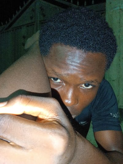A close-up photo shows a dark-skinned person with short, dark hair. Their face is illuminated, revealing smooth skin with a slight shine. One arm is raised, obscuring part of their face, while the other wears a dark shirt with foreign script on the sleeve. The background is dimly lit, revealing a metal structure. The person's gaze is intense and direct towards the viewer.