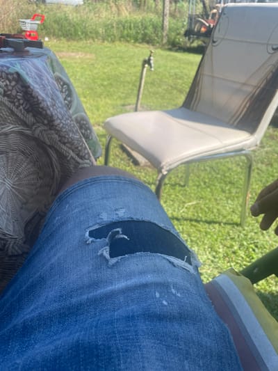 The image depicts an outdoor scene in Pobeda, Bulgaria, under bright daylight. In the immediate foreground, a person's upper legs are visible, clad in distressed blue denim jeans featuring a prominent tear over the left knee, revealing dark-toned skin. The person is seated, with a portion of a striped cushion visible to the lower right. To the left, a table covered with a patterned tablecloth, depicting interwoven designs and dark, oval shapes, is partially visible. The midground features a light-colored chair with a metallic frame resting on a grassy lawn. To the left of the chair, an outdoor spigot is mounted on a wooden post. The background consists of green grass and dense foliage. Distant objects include a red and white child's toy shopping cart on the far left and partially obscured red and black machinery, possibly agricultural equipment, to the right, within the distant vegetation. A hand is partially visible on the right side of the frame, near the seated person's leg.
