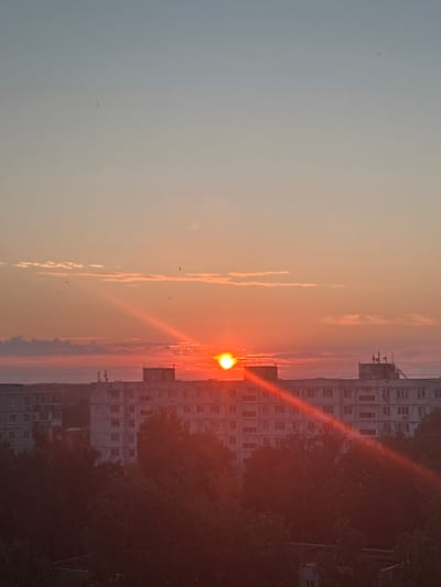 The image depicts a city skyline at sunset. The sun, a bright orange orb, is positioned just above the horizon, casting a warm glow across the sky. Rays of sunlight emanate from the sun, creating a lens flare effect that streaks across the frame. In the foreground, several apartment buildings rise above a dense canopy of trees. The buildings are several stories high and appear to be made of concrete. Some of the buildings have antennas on their roofs. The sky transitions from a soft blue at the top to a warm orange and pink hue near the horizon. A few wispy clouds are scattered across the sky. The overall impression is one of a peaceful evening in an urban setting.