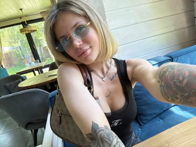 The image is a close-up, eye-level selfie of a young woman taken indoors, likely in a cafe or restaurant in Ozernoe, Russia, during daytime. The woman has short, straight blonde hair and blue eyes, partially obscured by light blue, rimless sunglasses with gold accents. She has a subtle smile and looks directly into the camera.

She is wearing a black ribbed tank top with a white cursive "G" or similar letter logo on the lower chest area. A silver-toned necklace with a heart-shaped pendant adorns her neck. Two tattoos are visible on her chest: one above her cleavage featuring cursive text, and another partially visible within the cleavage that appears to read "lure!".

Her left arm, held up to take the photo, displays a large, monochrome floral tattoo, primarily depicting roses. A brown handbag with a distinctive monogram pattern, suggestive of the Guess brand, hangs from her right shoulder. Another tattoo, a vibrant, colorful design with red and yellow tones, covers her right arm and shoulder. A simpler, short horizontal line tattoo is also visible on her right bicep.

The background reveals a bright, contemporary interior with light wooden plank walls and large windows offering a view of green trees and foliage under natural daylight, suggesting a sunny outdoor environment. Inside, there's a wooden table, dark grey upholstered chairs, and a blue cushioned sofa or bench along the wall. A round, wicker or natural material pendant lamp hangs from the ceiling. The overall atmosphere is casual and bright.