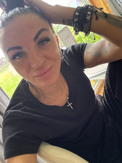 A close-up selfie shows a woman with dark hair pulled up in a bun, light blue eyes, and dramatic makeup. She is wearing a black t-shirt, a silver necklace with a cross pendant, and multiple bracelets on her left wrist. A nose ring is visible. Tattoos are present on her arms. She is indoors, near a window, with greenery visible outside. A small white box with text is also in the background. The setting appears to be a residential area.