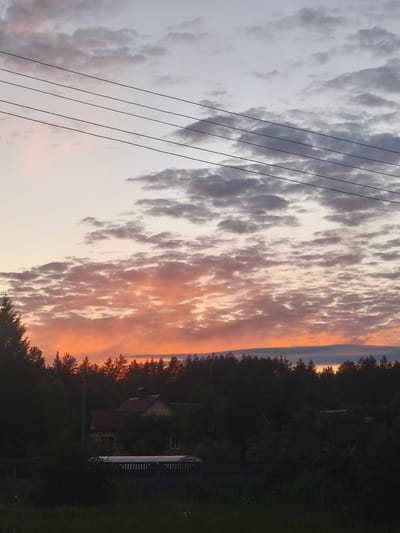 The image depicts a rural landscape at dusk, likely in or around Парэчча, Belarus. The sky is filled with dramatic clouds, transitioning from pale grey and lavender at the top to vibrant hues of orange and pink closer to the horizon. A dense line of dark green trees forms the background, silhouetted against the colorful sky. In the mid-ground, partially obscured by trees, is a yellow house with a red roof. In the foreground, there is dark vegetation and a wooden fence. Four parallel black wires stretch across the upper portion of the frame, suggesting the presence of overhead power lines. There are no people or animals visible in the scene. The overall impression is one of a quiet, serene evening in the countryside.