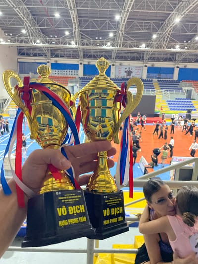 In a sporting arena, two gold trophies with red, white, and blue ribbons are held in the foreground by a person's hands. The trophies have black bases with yellow text that reads "VÔ ĐỊCH HANG PHONG TRÀO." In the background, a group of people in black and white attire are dancing on a red floor. Stadium seating, painted blue and orange, is visible behind the dancers, and a large screen displays images. The arena has a high, industrial ceiling with exposed metal beams and lighting fixtures. The location is identified as the city of Thuận An, Vietnam.