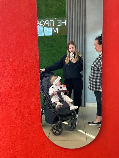 The image is a reflection in a mirror showing a woman with blonde hair and a black hoodie holding a smartphone and taking a selfie. In front of her, a baby wearing a white headband and a patterned outfit lies in a stroller. The woman appears to be looking at her phone, while the baby is looking slightly upwards.
The background of the reflection shows a wall with horizontal grey stripes and a green surface that resembles artificial grass, possibly a wall decoration or a green screen. A neon sign is visible, with some letters illuminated. The illuminated letters appear to spell "WEALTH" from left to right. The entire scene is framed by a textured red border.
Another reflection shows a person from the chest up, wearing a black and white checkered shirt and dark pants. This person's reflection is positioned above the woman and baby. The overall lighting suggests it might be daytime, though there are no strong indicators of the weather. The setting appears to be indoors, possibly a modern establishment or a studio, given the wall decorations and neon sign. The emotion conveyed is casual, perhaps a moment captured during an outing or a visit.
