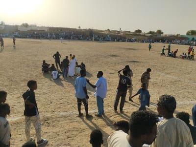 A dirt field hosts an event with gathered people. A crowd lines the perimeter with buildings in the background. Individuals on the field are casually dressed. On the right, players in green and yellow are gathered together. One individual stands alone in the distance. The lighting suggests the scene is during the day, possibly late afternoon.