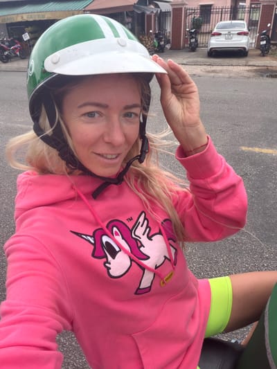 A blonde woman, wearing a green and white helmet, a pink hoodie with a unicorn design, and neon green shorts, is sitting on a vehicle, possibly a scooter or motorcycle, in an outdoor urban environment. Her left hand is on the brim of her helmet. In the background, there are buildings with tiled roofs, parked motorcycles, and a white car. The scene appears to be on a paved road. The context of Xuân Hương - Đà Lạt, Vietnam, suggests this is likely a street in that city.