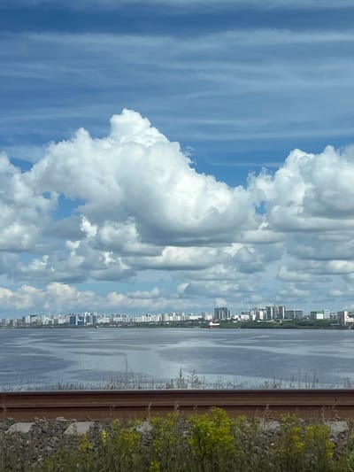 The image captures a wide, tranquil body of water, likely the Kama River, under a dynamic sky filled with large, white cumulus clouds against a bright blue backdrop. In the distance, a long, flat horizon is punctuated by a dense cityscape. The buildings appear to be primarily modern high-rise apartments and other urban structures, suggesting a developed area. The foreground is occupied by a section of railway track, with rusty brown rails resting on ballast. Beyond the tracks, a strip of green vegetation, including tall grasses and some yellow wildflowers, forms the immediate edge of the scene. The overall impression is one of a view from a vantage point near the riverbank, overlooking a city and its waterfront, with the infrastructure of a railway line in the foreground. This scene is consistent with the riverside urban landscape of Naberezhnye Chelny, Russia, a city situated on the Kama River and known for its extensive industrial and residential areas.