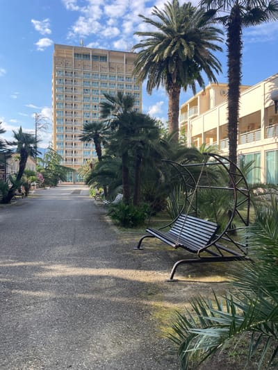 The image depicts an outdoor scene, likely a park or courtyard, with a paved pathway leading into the distance. Flanking the pathway are lush green palm trees of varying sizes and species. In the foreground to the right, a dark metal swing set with a slatted wooden bench is prominently featured. Further down the pathway, to the left, is a light-colored, multi-story building with numerous balconies. To the right of this building, a lower, lighter-colored structure with balconies and large windows is visible. The sky above is blue with scattered white clouds. The lighting suggests it is daytime, with sunlight casting shadows on the pathway. The environment appears to be well-maintained and tropical or subtropical in climate, consistent with a location like Gagra, Abkhazia. There are no people visible in the image.