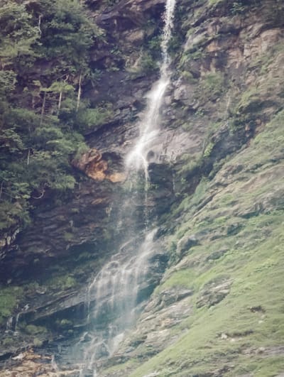 A waterfall cascades down a rocky mountainside covered in lush green vegetation. The waterfall is the dominant feature, a white ribbon of water against the dark, textured rock. The surrounding cliffs are steep and rugged, with patches of trees and moss clinging to their surfaces. The overall impression is one of natural beauty and rugged wilderness. The location in Kothi, India, suggests a mountainous region known for its scenic landscapes.