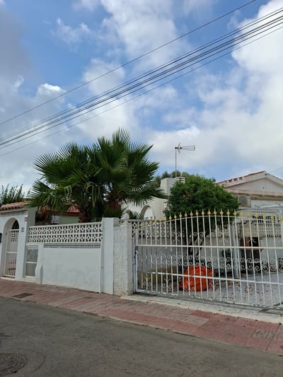 The image shows a residential street scene in Torrevieja, Spain. A white house with a tiled roof is visible behind a white metal fence with ornate gold finials and a white patterned wall. A large palm tree and a smaller green shrub are planted in front of the house. Power lines and a television antenna are visible against a cloudy blue sky. The street in front of the property is paved with asphalt, with a sidewalk made of red bricks and concrete tiles. An orange garbage bag is placed behind the gate. A manhole cover is visible on the street. There are no people visible in the image.