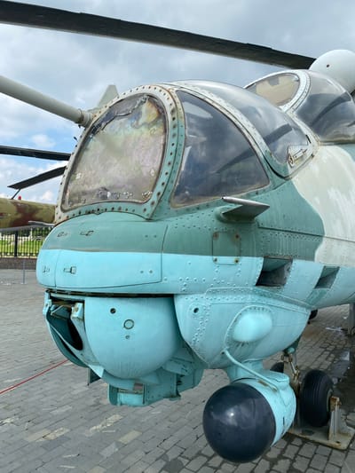 A close-up, eye-level, full shot captures the front portion of a faded teal and camouflage-patterned military helicopter. The helicopter is parked on a paved surface of gray and tan rectangular stones. The helicopter's canopy, made of multi-paneled, aged glass, is weathered and cracked, reflecting the overcast sky and surrounding environment. Beneath the canopy, on the nose of the helicopter, are two bulbous, teal-colored structures, suggesting a gun or sensor housing. A black spherical object, likely a sensor or munition, is visible protruding from the lower front section, supported by a landing gear strut with a wheel. In the background, another military helicopter, camouflaged in brown and green, is partially visible behind a metal fence. The scene is devoid of people, and the overall impression is one of a static display, likely in a museum or memorial site, such as the Military Technology Museum in Verkhnyaya Pyshma, Russia, which houses a collection of Soviet-era military hardware.