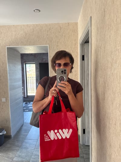 A woman is taking a selfie in a hallway. She is wearing sunglasses and a brown t-shirt and is holding a red shopping bag and a set of keys. The hallway has textured beige wallpaper and grey tiled floors. There is a mirror on the wall, and the woman is using her phone to take the picture. Behind her, there is a doorway and a window.