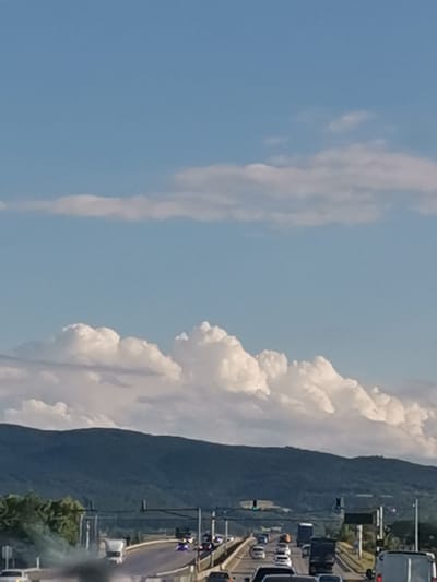 The image shows a highway with traffic, with a mountainous landscape and a cloudy sky in the background. The highway is multi-lane and there are various vehicles on it, including cars, trucks, and vans. Some vehicles are moving, while others appear to be stopped or moving slowly. There is a significant amount of dark smoke emanating from the left side of the image, obscuring some of the vehicles and the road. The sky is a bright blue with scattered white clouds. The mountains in the background are covered in green vegetation, suggesting a rural or natural setting. The scene appears to be during daylight. This view is consistent with the location in Kazichene, Bulgaria, known for its highway access and surrounding natural terrain.