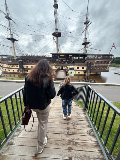 In Veliky Novgorod, Russia, a wooden staircase with a metal railing leads down towards a large, docked ship resembling a frigate, named "Фрегат Флагман" (Frigate Flagman). Two individuals, a woman with long dark hair and a child with lighter hair, are walking down the stairs. The woman is in the foreground, wearing a black leather jacket and beige pants, carrying a brown handbag. The child is in front of her, wearing a denim jacket and jeans. The ship is docked on a body of water with a cloudy sky overhead. The ship is a multi-story structure with masts and rigging, appearing to be a restaurant or museum. The environment is an outdoor setting with grass visible on the left side of the stairs.