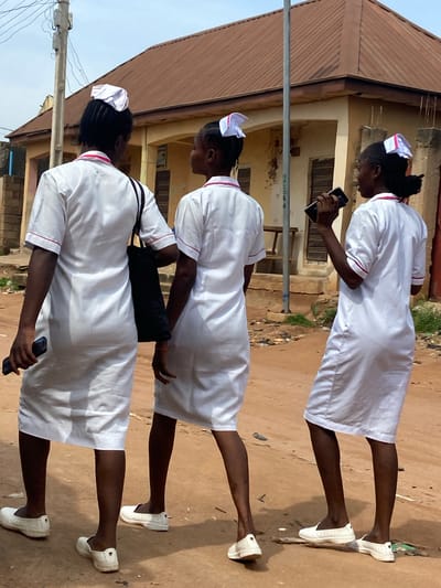 Three young women dressed in white nurse uniforms and white nurse caps are walking on a dirt path. The uniforms have thin pink trim around the neckline and cuffs. Each woman is wearing white lace-up sneakers. The first woman from the bottom is looking down at her phone. The second woman is looking down and to her right. The third woman is looking down at her phone. A black bag is slung over the shoulder of the second woman. They are walking in front of a weathered building with a corrugated metal roof. The scene appears to be outdoors during the daytime. There is no visible text in the image.