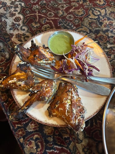 A top-down shot shows a plate of tandoori chicken, salad, and a small metal bowl of chutney, placed on a patterned fabric, likely a tablecloth or rug. The chicken pieces, visibly charred, are adorned with a white sauce or marinade. The salad consists of shredded cabbage and carrots. Silverware (fork and spoon) is placed atop the salad. The plate is a neutral color with a dark brown rim. The scene is indoors. There is no clear indication of the location being Arambol, India.