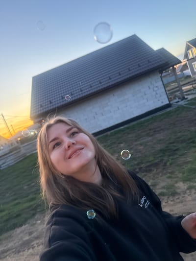 A young woman stands outside in the city of Ферма, Russia. She is wearing a black hooded sweatshirt. Several bubbles are floating in the air around her. In the background, there is a building with a dark roof and light-colored siding, and a grassy area. The sky is a pale blue-orange color.