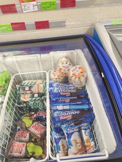 The image shows the interior of a commercial freezer display case filled with various ice cream products. The freezer is stocked with multiple types of packaged ice cream bars and tubs. In the foreground, a white wire basket contains several individually wrapped ice cream bars with brown and teal packaging, featuring the brand name "Jahis" and images of pears and chocolate. To the right of this basket, another white wire basket holds numerous individually wrapped ice cream bars in blue packaging, featuring "Белый Полюс" (White Pole) branding and illustrations of ice cream cones. In the upper right corner of the freezer, several clear plastic tubs of ice cream, also branded "Наше" (Our), are visible, some with fruit pieces. The freezer compartment has frosted edges. Above the freezer, price tags are attached to a shelf. Based on the presence of Cyrillic text and product branding, the scene is likely in a retail environment in Belarus. Given the store is located in Navapolatsk, Belarus, this image captures a segment of an aisle within a retail establishment in that city, specifically showcasing frozen dairy desserts for sale.