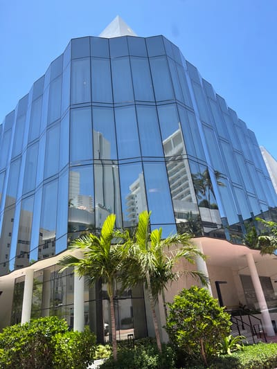 A low-angle, full-shot photograph captures a modern, multi-story building with a distinctive, angular facade clad in reflective blue glass. The building is situated in Miami Beach, Florida, as evidenced by the distinct architecture and lush tropical vegetation. The glass panels are arranged in a series of angled facets, creating a faceted, crystalline appearance. The building's upper section is topped with a sharp, pyramidal spire.
The reflections in the glass reveal the surrounding environment, showcasing other buildings, palm trees, and a clear blue sky. Among the reflections are high-rise residential buildings with white facades and balconies, characteristic of Miami Beach's coastal architecture.
In the foreground, a well-maintained landscape features various types of tropical greenery. Two prominent palm trees with feathery fronds stand closest to the viewer, their leaves catching the sunlight. Alongside them are dense bushes and smaller leafy plants, creating a vibrant green border.
The base of the building features a covered entrance area supported by white cylindrical columns. A set of stairs with black railings leads upwards to the right of the frame. The overall scene suggests a bright, sunny day in a vibrant urban setting with a strong tropical influence. There are no people visible in the image.