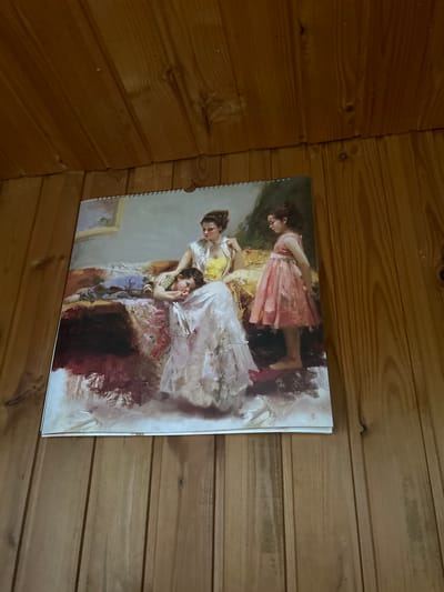 A painting hangs on a wooden paneled wall. The painting depicts a mother with two children. The mother is seated, wearing a white dress and a yellow bodice, and is holding a sleeping child on her lap. The child is wearing a blue jumpsuit and a striped shirt. To the right of the mother, a young girl stands wearing a pink dress. The background of the painting is a dimly lit interior scene, possibly a room. The wooden paneled wall suggests an indoor setting, possibly a home or cabin. The image does not contain any information about the city of Leningradskaya, Russia.