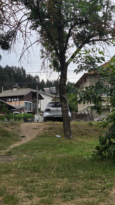 The image depicts a rural setting in Bania, Bulgaria, featuring several residential buildings, a car, and abundant vegetation. In the foreground, a dirt path winds through a grassy area with scattered wildflowers. A mature tree stands prominently in the mid-ground, its branches extending across the frame. Behind the tree, a silver Volkswagen Golf is parked.
To the left of the car is a two-story house with a brown wooden exterior, a gray tiled roof, and several windows. Two outdoor air conditioning units are visible on the side of this house. Adjacent to it, another house, painted white with a red-tiled roof, features solar water heaters on its roof. To the right of the parked car, a pile of cut firewood is stacked against a wall, and further to the right, another house with a light-colored facade and a red-tiled roof is partially visible. The background is dominated by a dense forest of evergreen trees. The sky is overcast, suggesting a cloudy day. The overall scene conveys a quiet, residential environment typical of a Bulgarian village.