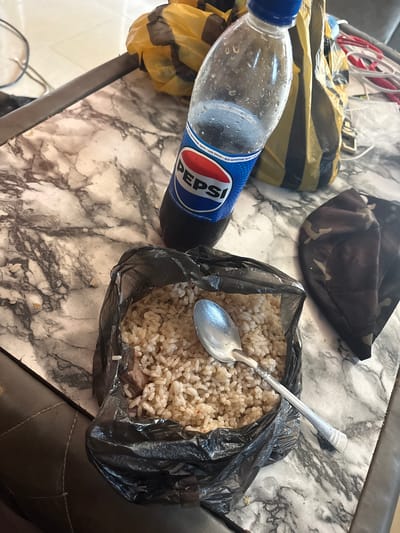 The image presents a casual meal setup on a light-colored table with a gray and black marble-like pattern. The primary subjects are a plastic bottle of Pepsi and a meal of rice served in a black plastic bag.

The clear plastic Pepsi bottle has a blue cap and a blue label. Visible text on the label includes "PEPSI," "COLA FLAVOURED CARBONATED SOFT DRINK," "NEW REFRESHING TASTE," and "LESS SUGARS." The bottle shows condensation, indicating it is cold.

The rice, which appears to be light-colored (possibly white or light brown), is contained within an open black plastic bag. A silver spoon is placed directly into the rice, suggesting it is ready to be eaten or is currently being consumed. At the bottom of the rice, some darker, chunky pieces, likely meat or stew, are visible. This method of serving food in a plastic bag is common for street food or informal meals, aligning with the provided location context of Ikot Ekpene, Nigeria.

In the background, partially visible on the table, are a yellow and black striped item (possibly another plastic bag or a part of a delivery container) and a camouflage-patterned fabric item, which could be a face mask or a piece of clothing. To the right of the meal, a dark, textured surface, possibly an armrest of a chair or another piece of furniture, can be seen. The overall scene depicts a simple and everyday meal.