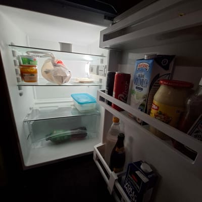 This media file shows the inside of a refrigerator in Rastatt, Germany, likely during daylight hours given the interior light. The refrigerator is moderately stocked with various food and drink items.
Prominent objects include several cartons of milk, specifically "Hofgut VOLLMILCH" and another dark blue carton labeled "VOLLMILCH" with "FAIRE HALTUNG" visible. There is also a red can of "River" soda and a dark glass bottle, possibly wine or beer, with a yellow cap. A glass jar with a red lid, labeled "PAR AMOUR DU GOUT," contains a light-colored spread. Other items include a blue plastic container, a clear plastic container holding a green bottle, and a plastic bag with a white handle containing food items. Smaller packages, one labeled "Bio," are also present on a lower shelf. The lighting within the refrigerator is illuminated by its internal light. The overall impression is of a typical home refrigerator interior, organized with various groceries.