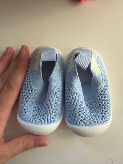 Two light blue knit baby shoes with white soles are positioned on a white surface. A human hand is touching the bottom shoe. The location cannot be determined from the visual information provided.