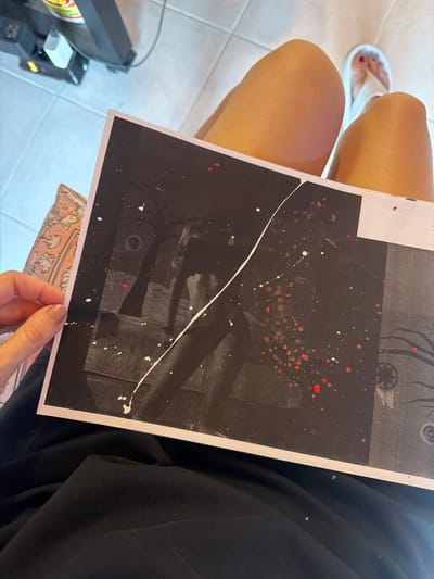A person's legs and feet are visible, sitting on a floor with white tiles. The person is wearing a black garment and white flip-flops. They are holding a piece of artwork, which is a black canvas with splatters of white and red paint. The artwork is positioned in front of the person's legs. In the background, a piece of furniture with electrical components and cables is visible. The scene appears to be indoors, likely in a home or studio. There is no discernible location within the city of Svilengrad, Bulgaria indicated in the image.