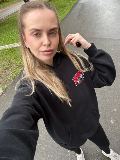 A full-body, top-down selfie of a blonde woman in Chernushka, Russia. She is wearing a black hoodie with a red and white logo on the left chest and black sweatpants. She has on white sneakers. Her hair is pulled back in a ponytail, and she is wearing light makeup. She is holding a black object in her right hand. The background is an asphalt path with grass on the left side. The lighting suggests it is daytime.
