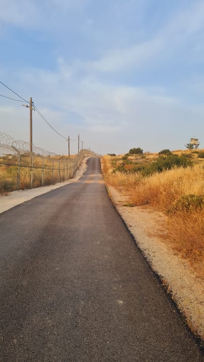 The image shows a narrow asphalt road cutting through a landscape. A fence runs along the left side of the road, reinforced with barbed wire. Wooden utility poles stand alongside the fence, with power lines visible against a blue sky. On the right side of the road, dry, golden vegetation covers the slope. In the distance, a watchtower stands on the hilltop. The road appears to stretch into the distance. The location is in the city of Lachish Regional Council, Israel.