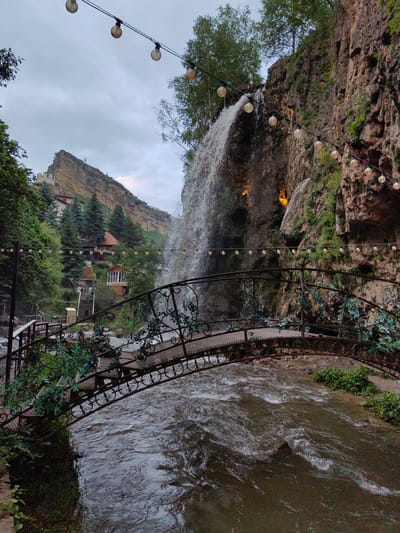 The image depicts a picturesque scene featuring a cascading waterfall flowing down a rocky cliff face, with a decorative metal bridge spanning a river in the foreground. The bridge is adorned with green vines and has an ornate, scrolled railing. String lights with round bulbs are suspended above the scene, illuminating the area with a warm glow. In the background, a rugged, multi-tiered mountain rises, with buildings nestled into its slopes, including a prominent reddish-brown roof structure. Lush green trees and vegetation are abundant throughout the landscape. The river below the bridge shows turbulent water with ripples and whitecaps. The sky is overcast, suggesting a cloudy or twilight condition. The location is identified as the city of Krasnyy Kurgan, Russia. No people are explicitly visible in the image, but the presence of the bridge and buildings suggests a developed area, possibly a park or tourist attraction.