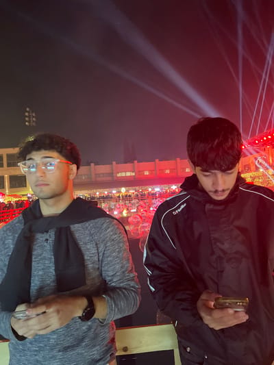 Two young men stand outdoors at night. The man in the grey shirt wears glasses and a dark scarf. He looks at the camera while holding a phone. Behind him is a lighted structure. The other man wears a black jacket with white stripes and looks down at his phone. Bright spotlights shine above the two men and buildings are visible.