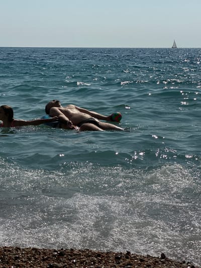 Two individuals are floating on a raft in the sea. The person on the right is a male, lying on his back with his arms outstretched. The person on the left appears to be a child, also in the water, and reaching out to the male. The water is a deep blue with shimmering sunlight reflecting off the surface. In the distance, a sailboat can be seen on the horizon, and the sky is clear and pale. The foreground of the image shows a rocky shoreline with a few waves breaking. The scene is set in Mirište, Montenegro.
