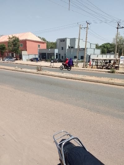 The image captures an outdoor scene from the perspective of someone seated on or immediately behind a motorcycle, looking forward down a paved road. The sky is clear and blue, indicating a sunny day, likely during late morning or early afternoon given the bright, direct light.
In the immediate foreground, the black padded seat and shiny silver metal luggage rack of a motorcycle are visible, extending into the bottom right corner of the frame.
The central part of the image is dominated by a wide, two-lane asphalt road, which appears to have a subtle median strip with sparse, dry vegetation separating the directions of traffic. On this road, in the mid-ground, a motorcycle is in motion, traveling from left to right. It carries two passengers; the driver is wearing a red top, and the passenger behind them is draped in a distinctive purple garment.
Beyond the road and median, on the other side, there are several buildings and activities. To the left, a two-story building with a reddish-orange pitched roof and lighter-colored walls stands out. It features multiple windows and appears to be a well-maintained structure. A few cars are parked in front of it, and some green trees partially obscure its lower levels.
To the right, a modern-looking, light grey building with a flat roof is prominent. This building has a commercial or institutional appearance, with what seems to be air conditioning units on its facade. A red sign or logo is visible on its side; while the text is difficult to fully decipher, it appears to contain the letter 'F' and possibly the word "Fidelity" or similar, judging by the shape. Several utility poles with a network of power lines crisscross the sky above this area. Adjacent to this building, further to the right, there is a rustic, open-sided wooden structure, possibly a market stall or shelter, with various items underneath it. Cars are also parked in front of this grey building.
In the background, several individuals can be seen walking or standing, adding to the sense of a populated area. The overall setting suggests a town or city, likely in a warm climate, given the dry vegetation and the style of architecture.