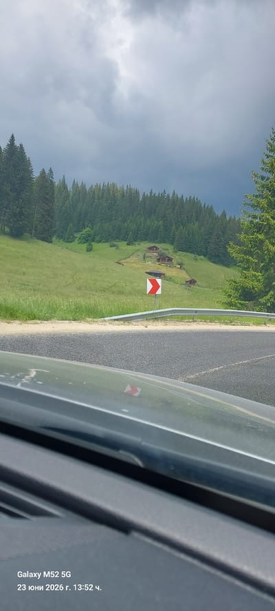 This is a landscape photograph taken from inside a vehicle. In the foreground, the dashboard of a dark-colored car is visible, reflecting a small red and white directional arrow sign. The middle ground features a paved road with a white line, bordered by a metal guardrail. Beyond the road, a grassy hillside rises, dotted with several small wooden houses and surrounded by dense evergreen forest. A bright red and white chevron sign is posted on the side of the road, indicating a turn. The sky is overcast with dark, dramatic clouds. The text overlay indicates the photo was taken with a "Galaxy M52 5G" on "23 юни 2026 г. 13:52 ч.", which translates to June 23, 2026, at 13:52. The location is identified as Smolyan, Bulgaria.
