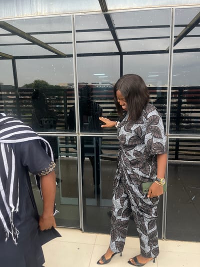 Two people are standing outside a building with large glass windows. The person in the foreground is wearing a black and white striped outfit with gold detailing on the sleeves. Their right arm is extended, holding a piece of black fabric. The second person, partially visible in the background, is wearing a patterned jumpsuit with a floral design in brown, black, and white. They are also wearing black high-heeled sandals and a watch on their left wrist. They are holding a green phone in their right hand and appear to be looking down at something. The scene is outdoors, with reflections of trees and buildings visible in the glass windows. The lighting suggests it is daytime.