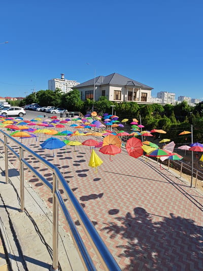 The image depicts an outdoor scene with a vibrant display of numerous colorful umbrellas suspended in the air, creating a visually striking installation. The umbrellas are of various colors, including red, orange, yellow, green, blue, purple, and pink, and are arranged at different heights, adding depth and dynamism to the scene. They are strung from wires or poles, forming a canopy over a pedestrian walkway paved with red and beige brickwork laid in a herringbone pattern.
In the background, a large, modern two-story house with a dark tiled roof and large windows is visible, situated on a slightly elevated area surrounded by lush green trees and bushes. Beyond the house, several multi-story apartment buildings are visible, indicating an urban or suburban setting. The sky is a clear, bright blue, suggesting a sunny day.
In the foreground, a silver metal railing runs along the left side of the image, bordering a concrete structure with visible cracks. The sunlight casts distinct shadows of the umbrellas and trees onto the paved ground, creating an interesting interplay of light and shadow.
To the left, several cars are parked on a paved surface, including a white SUV and other vehicles, suggesting proximity to a parking area.
The overall impression is one of an artistic public installation designed to add color and visual interest to a public space, likely a park, promenade, or a pedestrian zone in a city.
Based on external information, this installation of colorful umbrellas is located in the city of Aktau, Kazakhstan.
