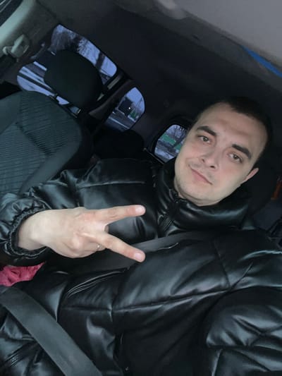 The image is a selfie of a man seated inside a car, likely in the front passenger seat. He has short dark hair, prominent dark eyebrows, and light brown eyes, with light stubble on his face. He wears a black, shiny puffer jacket and has a calm, slightly smiling expression while looking directly at the camera. His right hand is raised, forming a peace sign (V-sign) with two fingers, and a simple ring is visible on his right ring finger.
The interior of the car is visible, including a dark seatbelt across his chest and the textured fabric of the car seat to his left. Through the car windows, the exterior appears dimly lit, suggesting dusk, night, or a heavily overcast day. Patches of snow or frost are visible on the ground outside, indicating cold weather conditions. The overall mood conveyed by the man's expression and gesture is relaxed and positive. No visible text is present in the image. The location context provided is Novosinkovo, Russia, which aligns with the observed cold-weather environment.