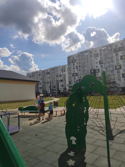 This image depicts an outdoor playground scene with several children engaged in play. In the background, a tall, modern apartment building dominates the skyline, with rows of windows and a light-colored facade. The sky is a vibrant blue, dotted with fluffy white clouds, indicating a sunny day, likely mid-morning or afternoon given the bright sunlight casting distinct shadows.
The playground itself is equipped with a green climbing structure shaped like an alligator, and a purple merry-go-round is partially visible in the foreground. Children of various ages are scattered across the playground. One child, wearing a blue shirt and hat, stands near the alligator structure, while another, in a red shirt and dark shorts, is interacting with a child in a pink shirt. A fourth child, also in a blue shirt and hat, is standing further back near the apartment building. They all appear to be enjoying themselves in a relaxed, playful manner. The ground surface of the playground is made of interlocking rubber tiles, typical for safety surfacing. The presence of the apartment building and the visible playground equipment suggests a residential area, likely a courtyard or public space within a town or city. The overall atmosphere is one of a pleasant, active summer day. No visible text is present in the image.