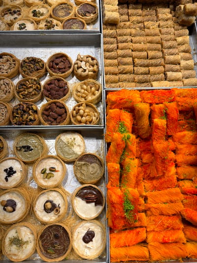 This close-up, overhead image showcases a vibrant and diverse array of Middle Eastern sweets meticulously arranged in metal trays, likely displayed in a bakery or sweet shop in Jerusalem, Israel.
In the top left tray, numerous golden-brown, cylindrical pastries are neatly packed in rows. These appear to be made from tightly rolled shredded phyllo dough, characteristic of kadaif-based sweets like 'Burma' or 'Kol W Shkor', often filled with nuts.
Adjacent to this, in the top right tray, are bright orange, rectangular pieces of dessert. These sweets are generously coated in syrup and sprinkled with crushed green pistachios, a distinctive presentation often associated with certain varieties of Knafeh, known for its vibrant orange color.
The bottom half of the image features two trays filled with smaller, individual round pastries, each nestled in a delicate cup or 'nest' made from shredded phyllo dough.
The bottom left tray primarily displays these nests filled with various whole nuts, including cashews, almonds, pistachios, and possibly pine nuts, each type typically occupying a separate basket-like portion.
The bottom right tray presents similar phyllo nests but filled with creamy or pudding-like substances. These fillings vary in color and topping; some are white cream topped with pistachios, pecans, or dried fruits like raisins, while others feature chocolate or a greenish cream, likely pistachio-flavored, adorned with different nuts or chocolate pieces.
The sweets are brightly lit, highlighting their intricate textures and appealing colors. No people are visible, and there is no discernible text in the image. The meticulous arrangement suggests a commercial setting designed to showcase the variety and freshness of the products.