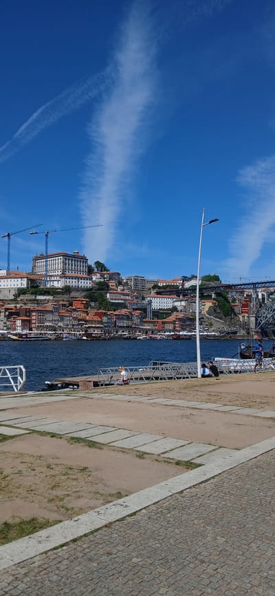 The image depicts a waterfront scene in Vila Nova de Gaia, Portugal, showcasing a section of the Douro River and a view across to the cityscape of Porto on the opposite bank. The sky is a bright blue, marked by streaks of white clouds.
The Douro River fills the center of the frame, reflecting the buildings along the Porto shoreline. The river separates the foreground from the background, where dense urban development is visible, characterized by a cluster of buildings with terra cotta roofs. Construction cranes are visible.
The foreground features a waterfront promenade or walkway, defined by a pavement surface and a metal railing that separates the walkway from the river. People are visible walking along the walkway.