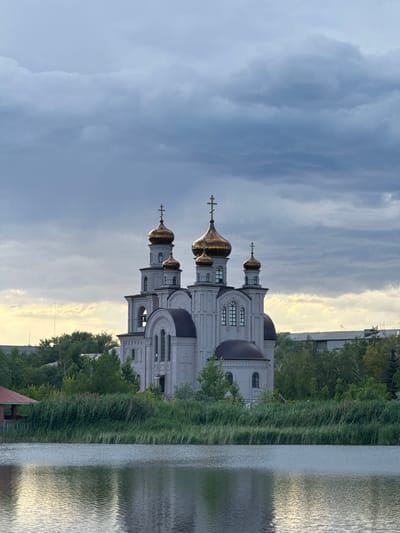 This media file depicts an Orthodox church situated beside a body of water, likely a river or lake, in Pavlodar, Kazakhstan. The church is a prominent structure with multiple domes, some of which are golden and reflect the light. It features traditional architectural elements such as arched windows and decorative molding. Surrounding the church are lush green trees and tall reeds growing along the water's edge. The water itself is calm, with ripples creating a textured surface that reflects the sky and surrounding greenery.
The sky is partly cloudy, with dramatic cloud formations suggesting either an approaching storm or the aftermath of one. The lighting indicates it is likely late afternoon or early evening, given the soft, diffused sunlight and the warm tones in the sky, particularly on the right side of the frame. There is no visible text in the image. The scene evokes a sense of tranquility and spiritual contemplation, enhanced by the natural beauty of the setting and the imposing presence of the church. The overall impression is one of a peaceful, perhaps slightly somber, moment in time.