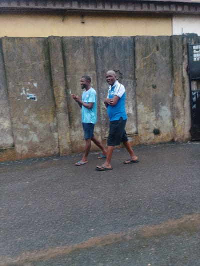 The scene depicts two adult males walking side-by-side on a damp asphalt road in the city of Agani, Nigeria. Behind them is a weathered, segmented concrete wall, topped by a section of a yellow-painted building. To the far right, a dark gate with an illegible white sign marked "GATE" is partially visible.
The male on the left, with a short beard, wears a light blue short-sleeved t-shirt, dark shorts, and light blue flip-flops. His hands are raised in front of him, palms slightly open, and he is smiling, looking to his right. The male on the right, bald, wears a blue and white collared polo shirt with a crest on the left chest, black shorts, and black flip-flops. His arms are crossed over his chest, and he is smiling while looking directly towards the observer. Both individuals are in mid-stride, moving from left to right across the frame.
