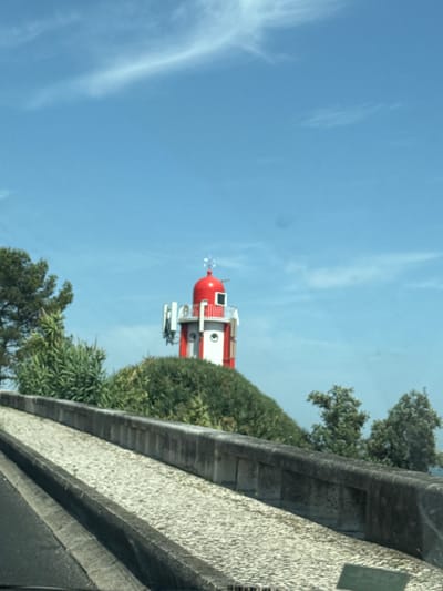 The image shows a red and white lighthouse situated on a grassy hill, under a clear blue sky with wispy clouds. The lighthouse has a rounded red roof and white cylindrical body with a red band. There are white antenna structures attached to the side of the lighthouse. The foreground features a stone wall and a cobblestone path, suggesting an elevated viewpoint. Lush green trees and vegetation surround the lighthouse. The scene appears to be in Oeiras, Portugal, possibly near the coast given the bright sky and open view. There are no people visible in the image. The overall impression is a clear, sunny day with a focus on the lighthouse structure and its natural surroundings.