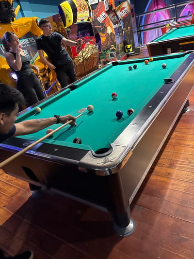 The image shows a billiards table in an entertainment establishment. The table is green and has several billiard balls on it. Two people are standing around the table, one of whom is holding a cue stick and appears to be aiming at a ball. Arcade machines are visible in the background. The specific location in Villa General Belgrano, Argentina, cannot be determined from the image alone.