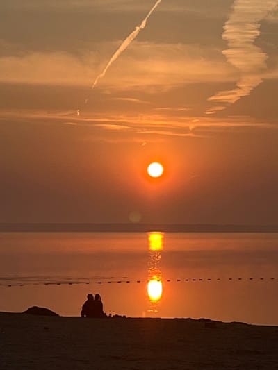 The image depicts a sunset over a body of water, likely a lake or sea, at a location in Surakovo, Russia. The sun, a bright orange orb, is positioned near the center of the frame, casting a warm glow across the sky and water. Its reflection creates a shimmering path on the calm surface. In the foreground, silhouetted figures of two people are seated on a sandy beach, facing the sunset. A dark, amorphous object, possibly a rock or piece of driftwood, is to their left. The sky is a gradient of orange and muted tones, with wispy clouds and contrails from an aircraft streaking across the upper portion. A line of buoys or markers extends horizontally across the water in the middle ground. The overall atmosphere is serene and peaceful.